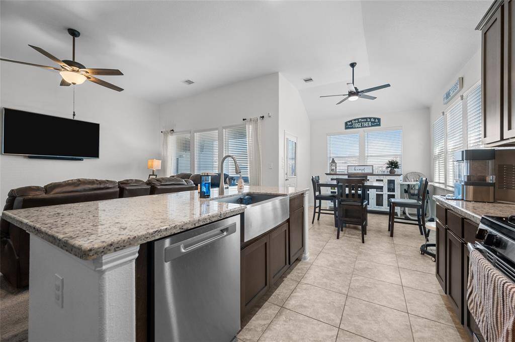 The kitchen features a large granite island, stainless steel appliances, and dark wood cabinetry. The adjoining living space includes comfortable seating and a mounted TV. Large windows provide ample natural light, and ceiling fans add comfort and style.