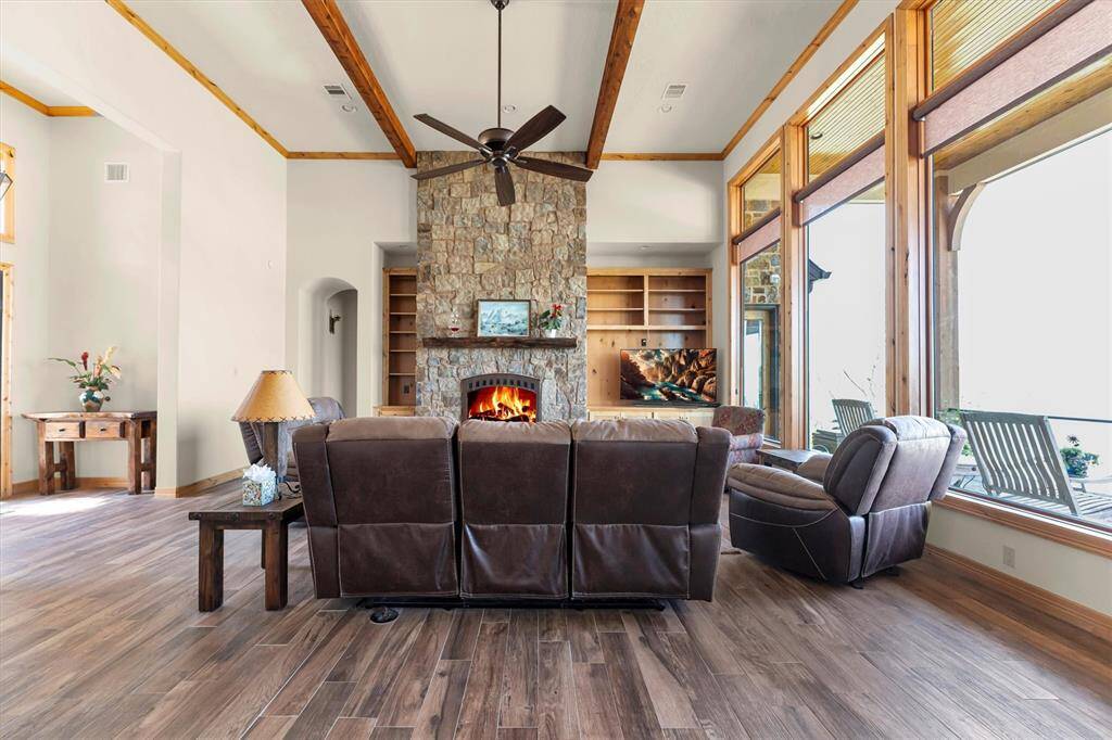 This stunning family room features wood-look tile flooring, a dramatic floor-to-ceiling stone fireplace, and a soaring ceiling with rich wood-beamed ceilings. Stylish built-ins add functionality while expansive picture windows frame captivating views of the patio, sparkling pool, and the lake.
