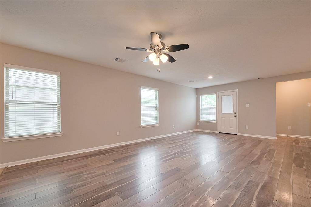 This spacious living area features light wood flooring, neutral walls, and ample natural light from multiple windows. A ceiling fan is centrally located, and there's convenient access to the outdoors through a door at the back.