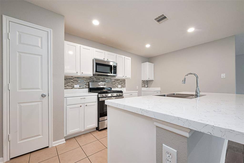 This modern kitchen features sleek white cabinetry, a stylish tile backsplash, and stainless steel appliances, including a gas stove and microwave. The spacious island with a quartz countertop and sink offers ample workspace and seating possibilities, while recessed lighting brightens the space.