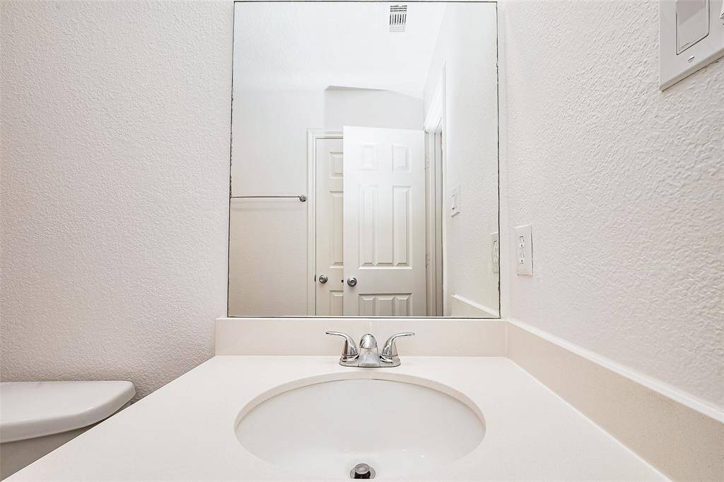 The photo features a clean, modern bathroom with a white countertop and an integrated sink. There's a large mirror above, a chrome faucet, and visible door and light switch. The space has a fresh, minimalistic design.