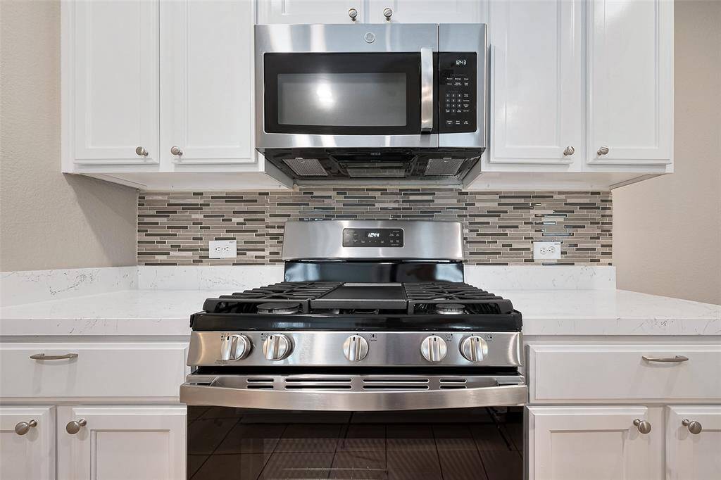 This modern kitchen features a stainless steel gas stove and microwave, set against a stylish tile backsplash. White cabinets and marble countertops add a sleek, contemporary touch.