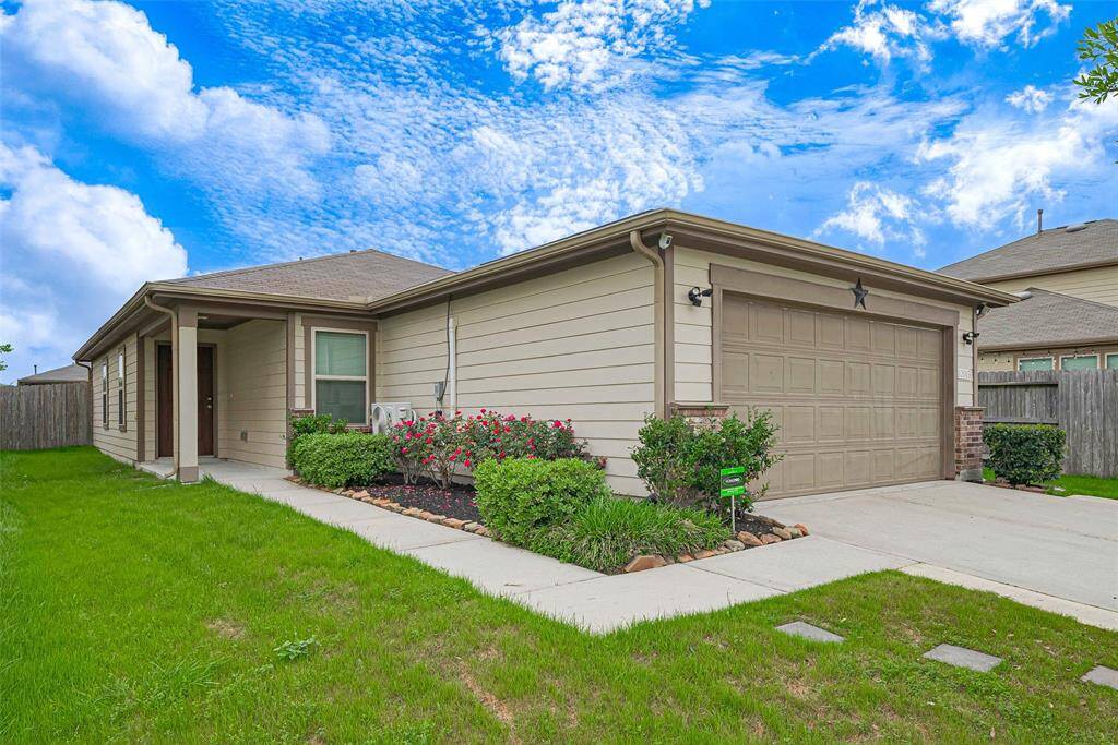 This photo depicts a charming single-story home with a two-car garage, a well-maintained front yard, and a covered entryway. It's surrounded by a wooden fence and features a bright exterior under a clear blue sky.
