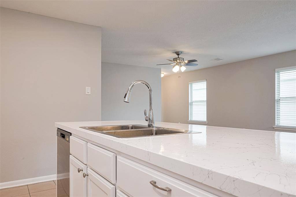 This photo showcases a modern kitchen with a sleek white quartz countertop, undermount sink, and stainless steel faucet. The open layout includes a view of a living area with natural light from multiple windows and a ceiling fan for comfort.