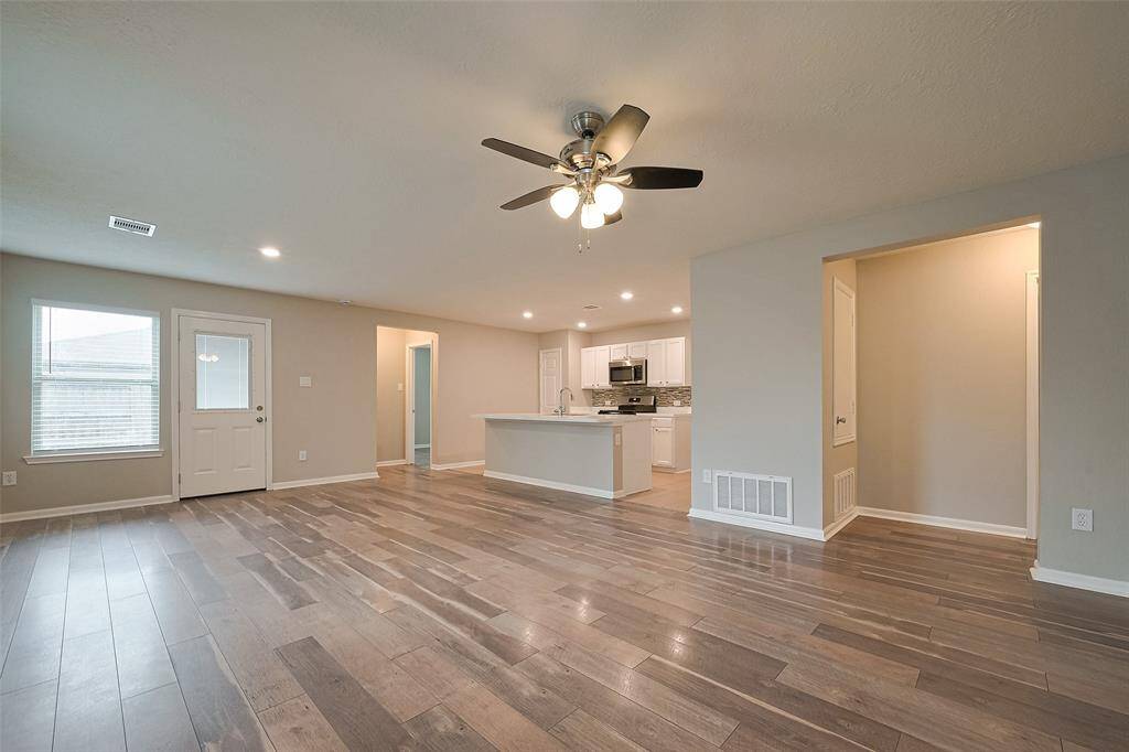 This spacious living area features modern wood flooring, neutral walls, and a ceiling fan. It seamlessly connects to a bright kitchen with white cabinetry and stainless steel appliances, offering a welcoming and open layout perfect for entertaining.