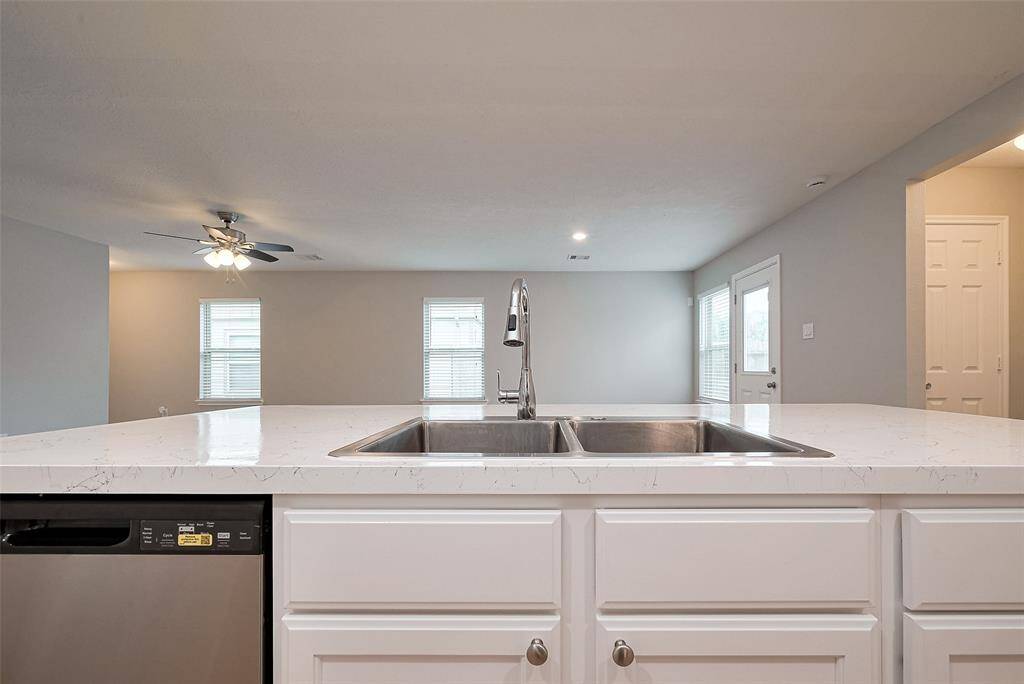 This photo showcases a modern, open-concept kitchen with a large marble-topped island featuring a double sink and stainless steel dishwasher. The space flows into a bright living area with ample natural light, ceiling fan, and access to a patio through a glass door.