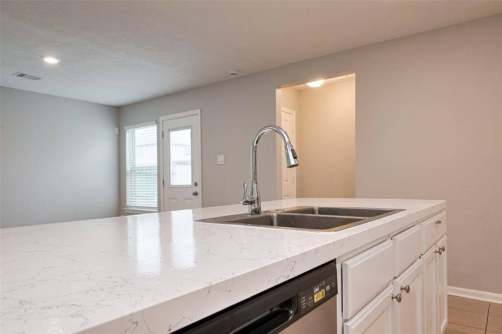 This photo showcases a modern kitchen with a sleek, spacious island featuring a marble-like countertop and stainless steel sink. The room is bright with neutral tones, recessed lighting, and a view of a door and window leading outside.