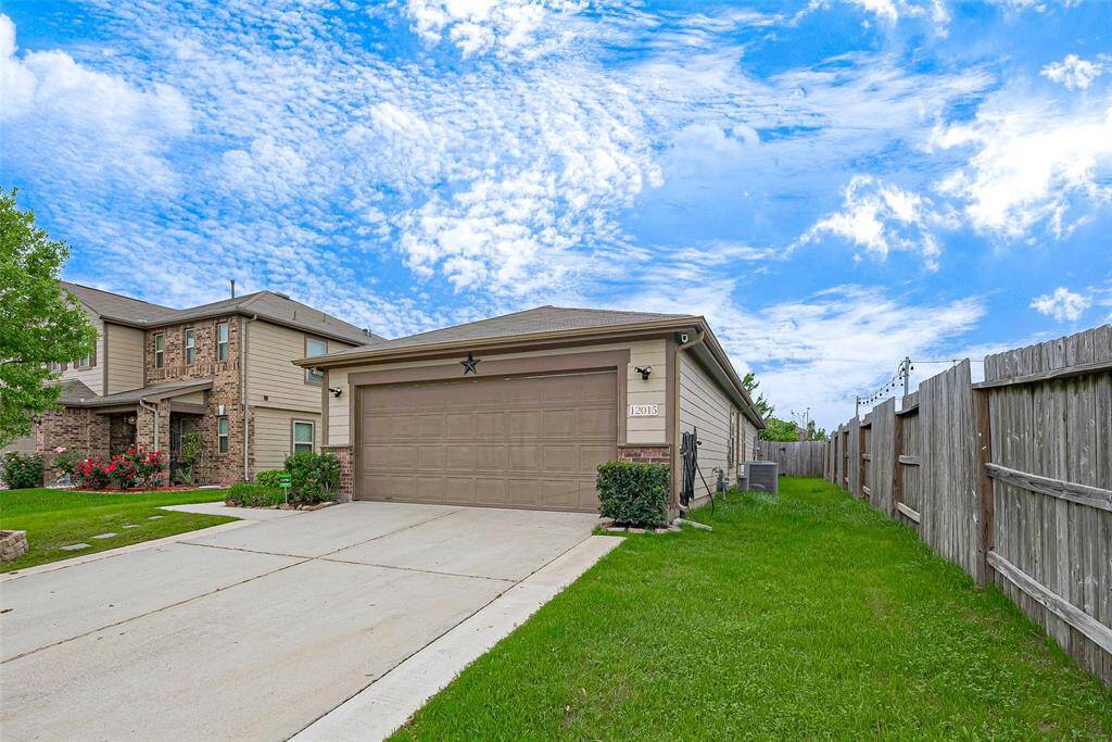 This photo shows a modern two-story home with a spacious driveway and attached garage. The exterior features brick and siding, with a well-maintained lawn and flowerbeds. A wooden fence encloses the side yard, and the sky is clear and blue.