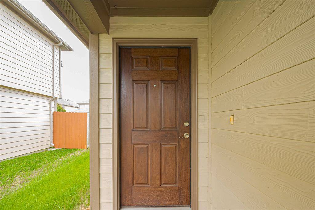 This photo shows a home's entrance with a solid wooden front door, surrounded by beige siding. The area is partially covered, and there's a glimpse of a neighboring house and a small grassy area with a wooden fence.
