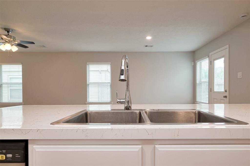 This photo showcases a modern kitchen with a sleek dual sink and a stylish faucet set in a bright, open layout. The space features a ceiling fan and ample natural light from large windows and a glass door, enhancing the airy feel.