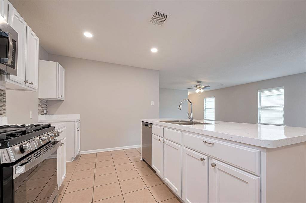 This photo showcases a modern kitchen with white cabinetry, stainless steel appliances, a spacious island with a sink, and tile flooring. The open layout leads into a well-lit living area with a ceiling fan.