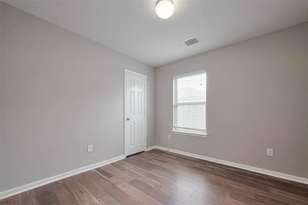 This room features neutral-colored walls, a window allowing natural light, and hardwood flooring. A simple light fixture and a door add to its functionality, making it ideal for a bedroom or office space.