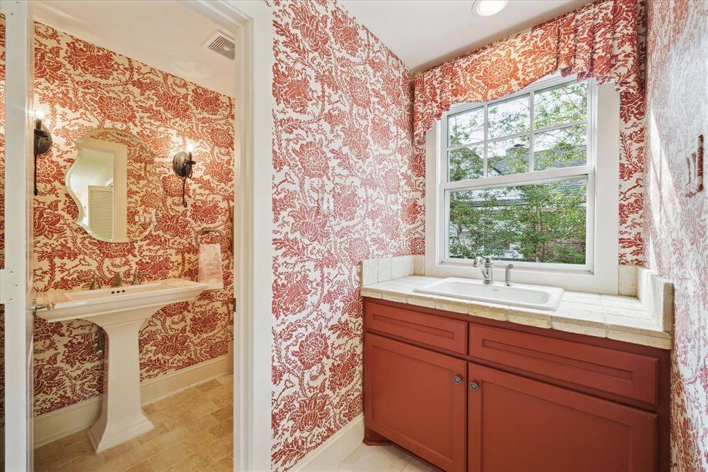 This powder bathroom off of the mudroom features vibrant red toile wallpaper and a white pedestal sink. The window offers a view of greenery, and the counter features a built-in sink. The space   combines classic and bold design elements with functionality.