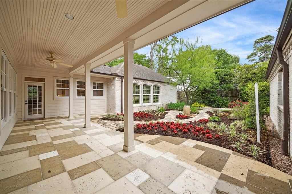 The expanded covered patio showcases tumbled travertine flooring, ceiling fans, and a view of a beautifully landscaped yard. The lovely flowers and lush greenery create a serene outdoor retreat that is seamlessly connected with the house.