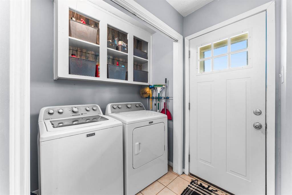 Well-organized laundry nook featuring a modern Maytag washer and dryer, built-in shelving with storage baskets, and convenient access to the backyard or garage.