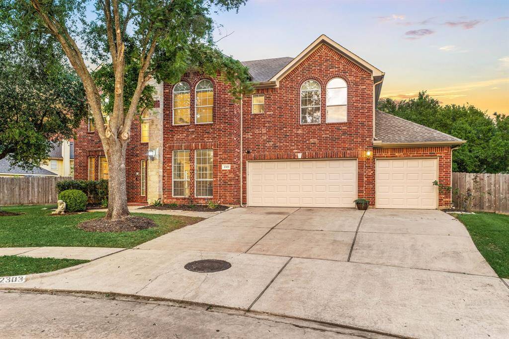 This is a spacious two-story brick home with a three-car garage, featuring large arched windows and a well-maintained front yard with mature trees. It sits on a wide driveway in a quiet neighborhood setting.