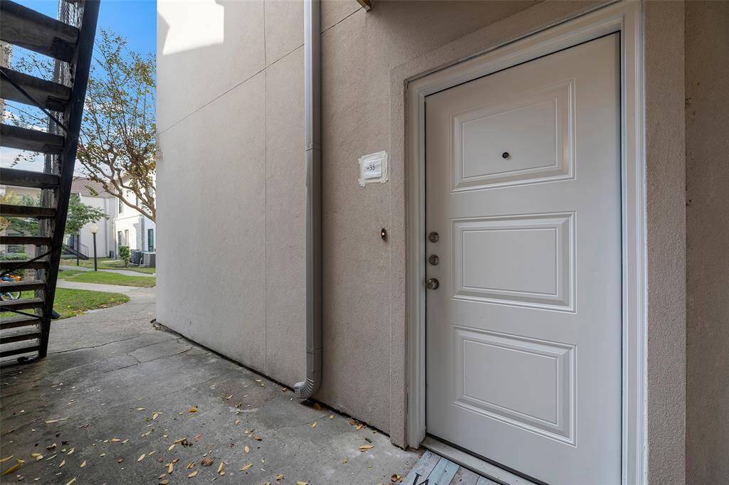 This photo shows the entrance to a ground-level unit, featuring a clean, white door with a peephole. The area is paved, with nearby stairs and a small lawn visible, suggesting a multi-unit residential setting.