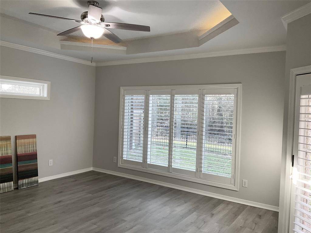 This room features light gray flooring, neutral walls, and large windows with plantation shutters, providing ample natural light. A ceiling fan with a light fixture is centered in the room, and a tray ceiling adds architectural interest.