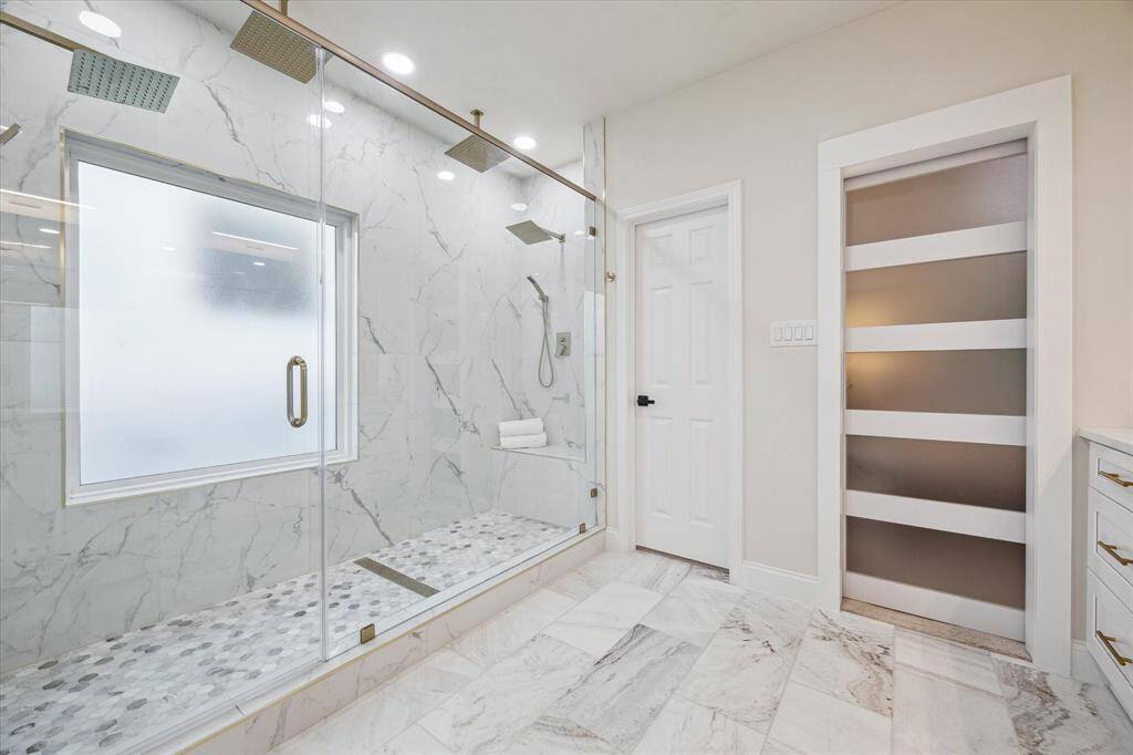 The primary bath retreat features a stunning extra-wide, oversized shower designed for ultimate relaxation and functionality. Dual shower heads on each side provide a spa-like experience, while the luxurious white marble walls create a clean, elegant backdrop. The linear drains add a sleek, modern touch, enhancing both style and practicality. A built-in seating bench offers comfort and convenience, and the rain shower heads provide a soothing, rainfall-like shower experience.