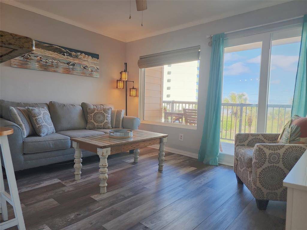 This cozy living room features a soothing color palette, modern ceiling fan, and stylish decor. It has a large window and sliding glass door leading to a balcony with a view of palm trees, creating a relaxing atmosphere.