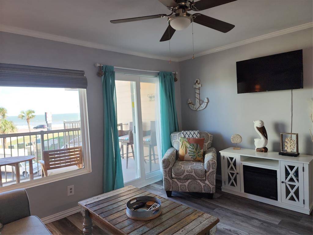 Cozy beach-themed living room with ocean views, featuring a comfy sofa, accent chair, ceiling fan, and sliding glass doors leading to a balcony. Light wood flooring and tasteful decor enhance the inviting atmosphere.
