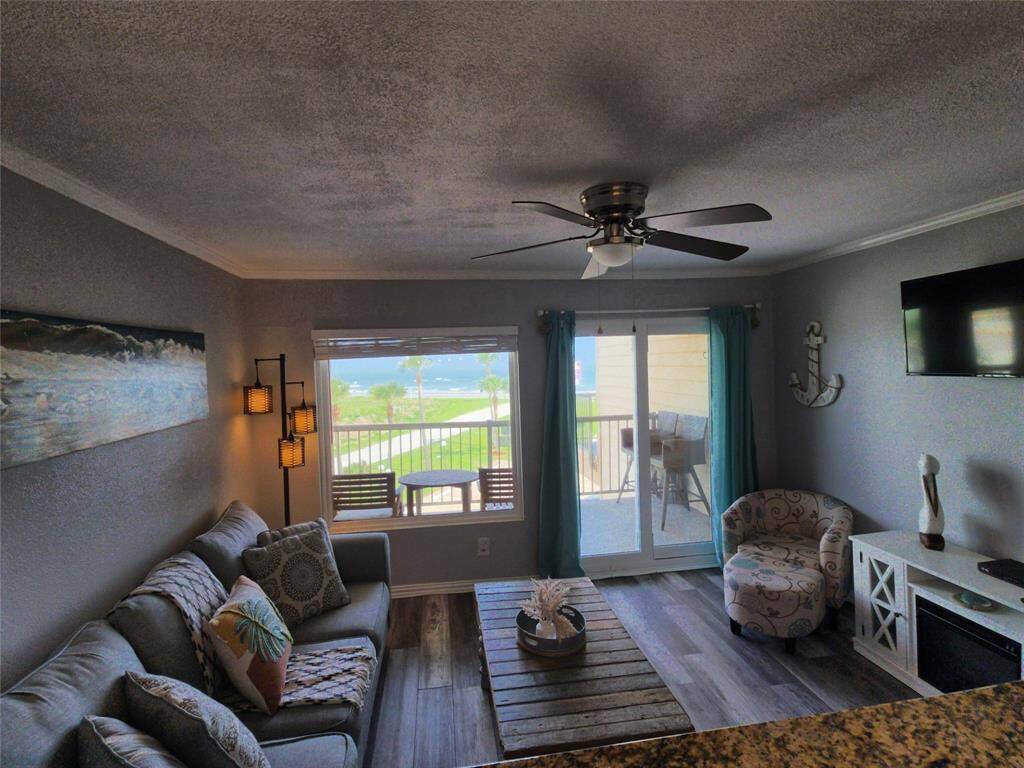 This cozy living area features a comfortable gray sofa with decorative pillows, a rustic wooden coffee table, and a stylish floor lamp. The open kitchen includes a granite countertop with bar seating and modern lighting. Art and decor add a warm touch to the space.