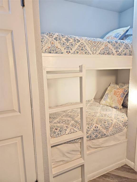 Cozy bedroom with a beach-themed decor, featuring a comfortable bed, ceiling fan, and warm lighting. The room has a coastal vibe with seashell-patterned bedding, an ocean painting, and decorative fish wall art. Natural light filters through orange curtains, creating a welcoming atmosphere.