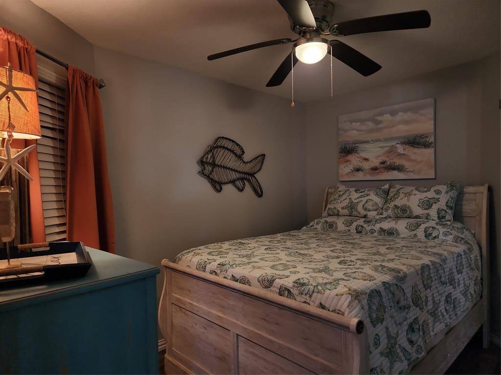 This cozy bedroom features a seashell-themed bedspread, a blue dresser, and coral-colored curtains. A ceiling fan provides comfort, and there's a wall-mounted fish decor. Adjacent is a room with bunk beds, adding extra sleeping space.