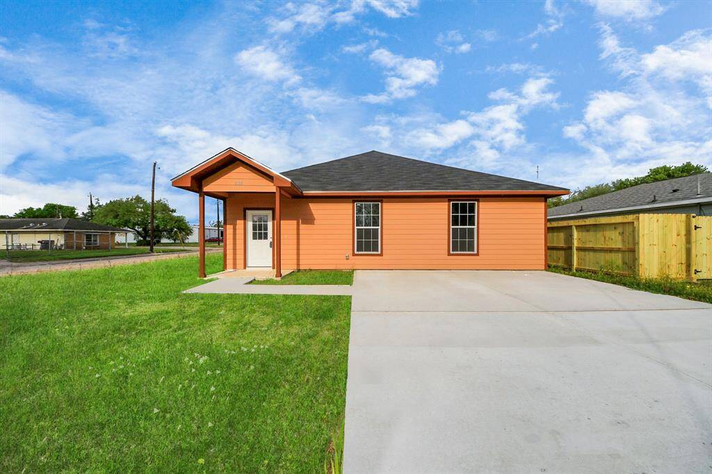This photo shows a single-story, orange house with a covered front porch and a black roof. It features a spacious concrete driveway and is set on a grassy lot. The property is bordered by a wooden fence, with a blue sky in the background.