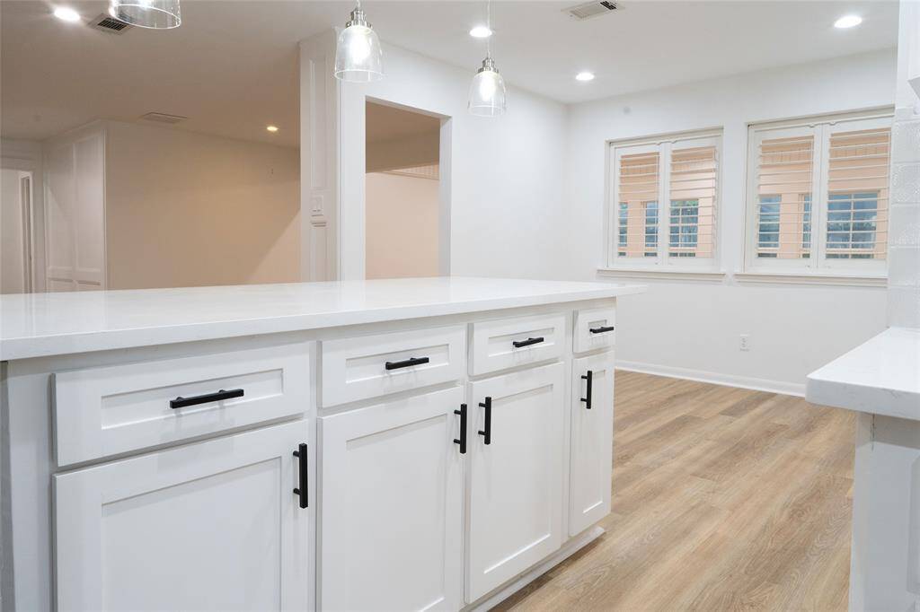 This modern kitchen features a spacious island with sleek white cabinetry, black hardware, and pendant lighting. The open layout is complemented by light LVP flooring and large windows with shutters, creating a bright and inviting space.