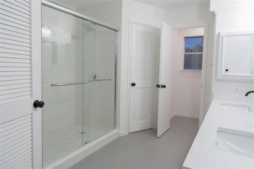 This bathroom features a spacious glass-enclosed shower, dual-sink vanity, sleek tile flooring, and a window for natural light, offering a clean and modern design.