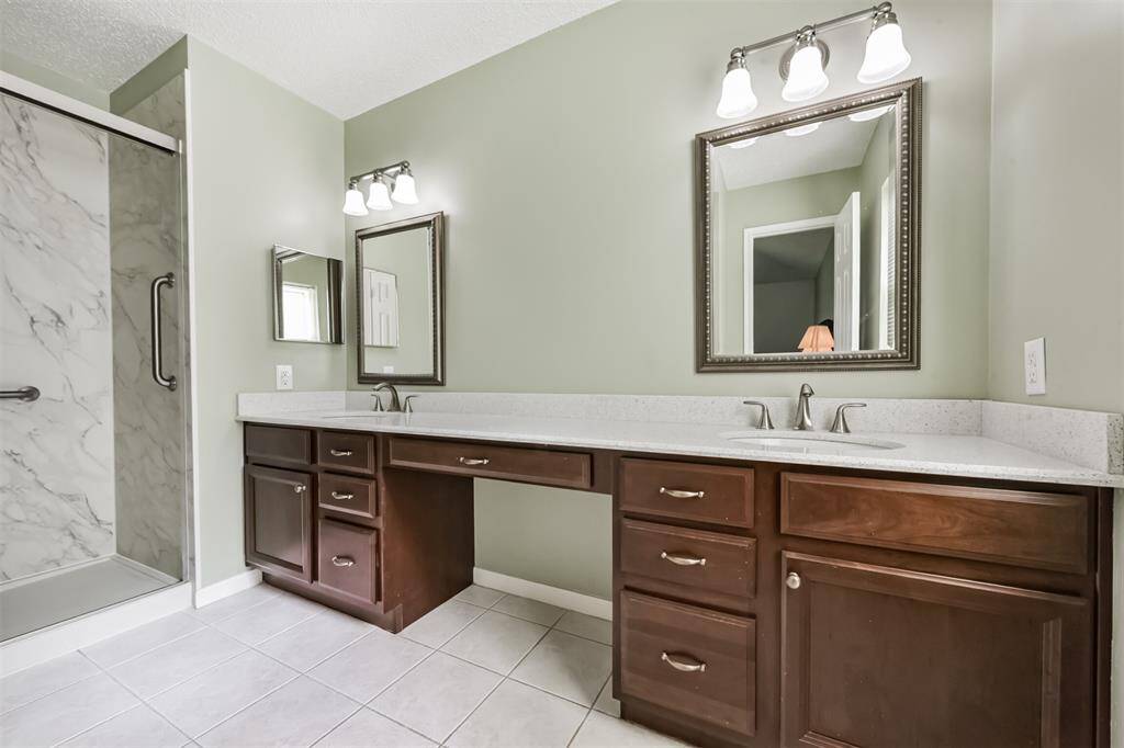 This bathroom features a dual-sink vanity with elegant mirrors and ample cabinetry. It has a spacious walk-in shower with a marble-like finish, complemented by soft green walls and tiled flooring.