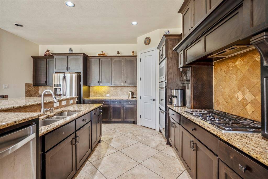 The chef-inspired kitchen features extensive cabinet storage, a striking tumbled stone backsplash, stainless steel appliances including double ovens, and a 5-burner gas cooktop. Granite countertops and under-cabinet lighting elevate the space with both elegance and function.