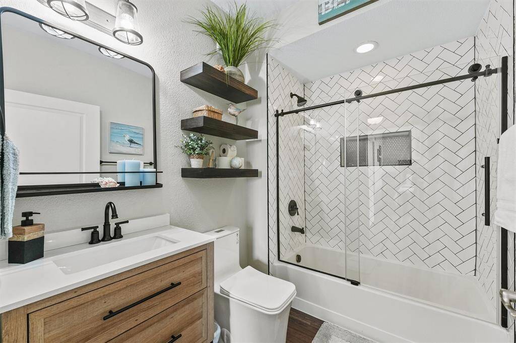 The guest bathroom has been beautifully updated with a chic vanity, black fixtures, floating shelves, and a herringbone-tiled tub/shower combo with sliding glass doors.