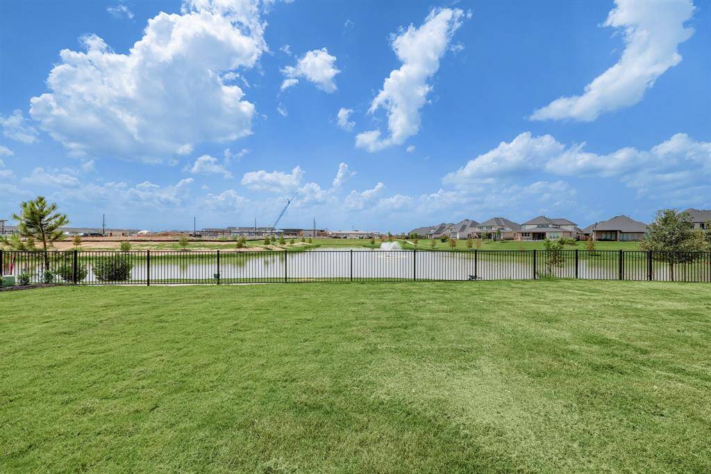 A spacious backyard with a well-maintained lawn overlooks a serene lake, surrounded by a wrought iron fence. The view includes nearby houses and a clear, blue sky, offering a peaceful and scenic setting.