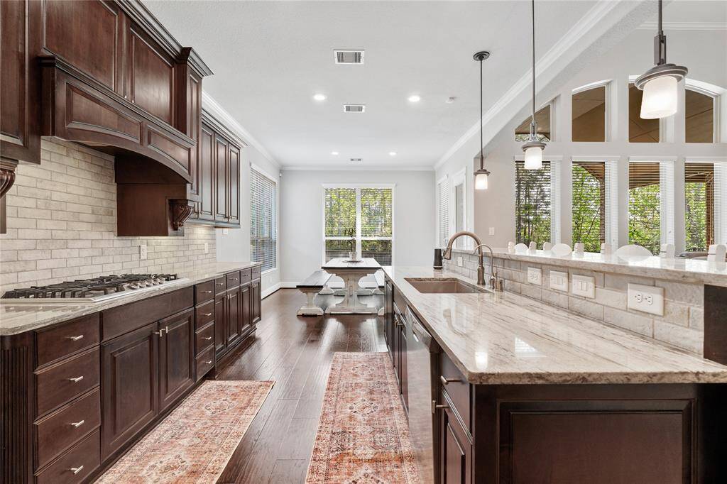 This spacious kitchen features elegant dark wood cabinetry, a large island with granite countertops, modern pendant lighting, and a gas cooktop. The open layout flows into a bright dining area with large windows, offering plenty of natural light and a view of the greenery outside.