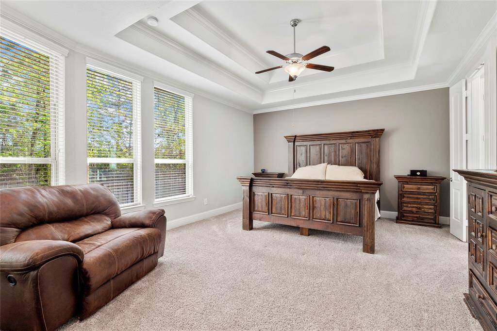 Primary bedroom is carpeted, has large windows providing natural light, a ceiling fan, and tray ceilings.