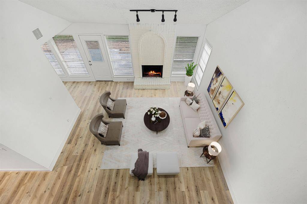 This spacious, open-concept living area boasts high ceilings and abundant natural light from large windows, offering a bright and airy atmosphere. The focal point is a beautifully textured white brick fireplace that adds warmth and charm, while the elegant wood-look flooring provides a sleek and modern foundation for versatile styling.