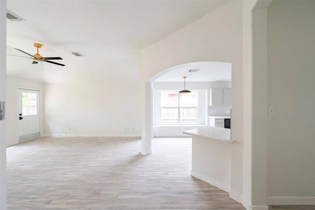 Bright, open-concept living space with white oak laminate wood flooring, a modern ceiling fan, and large windows offering natural light. The area seamlessly connects to a kitchen with a breakfast bar, enhancing the airy ambiance.