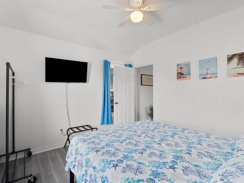 This bright bedroom features a ceiling fan, wall-mounted TV, and a minimalist clothing rack. The space is accented with beach-themed decor and vibrant blue curtains, creating a relaxed atmosphere.