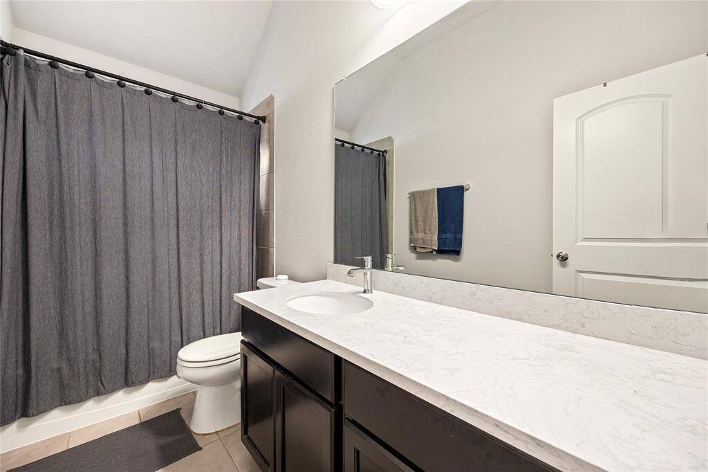 Secondary bath features tile flooring, bath/shower combo with tile surround, dark wood cabinets, beautiful light countertops, mirror, dark, sleek fixtures and modern finishes.