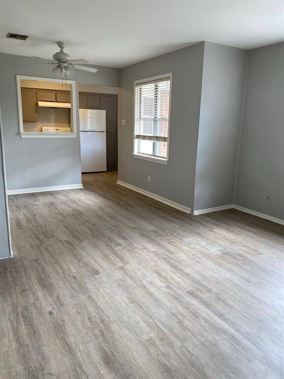 Bright, open living area with light wood flooring, neutral gray walls, and a ceiling fan. There's a window letting in natural light.