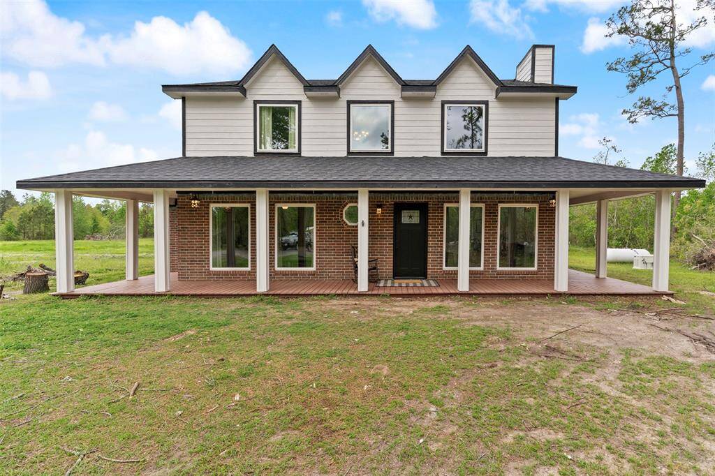 This is a charming two-story home with a brick and siding exterior. It features a welcoming front porch with a spacious overhang supported by white columns. The property is surrounded by a large, grassy yard with trees in the background, offering a serene, natural setting.