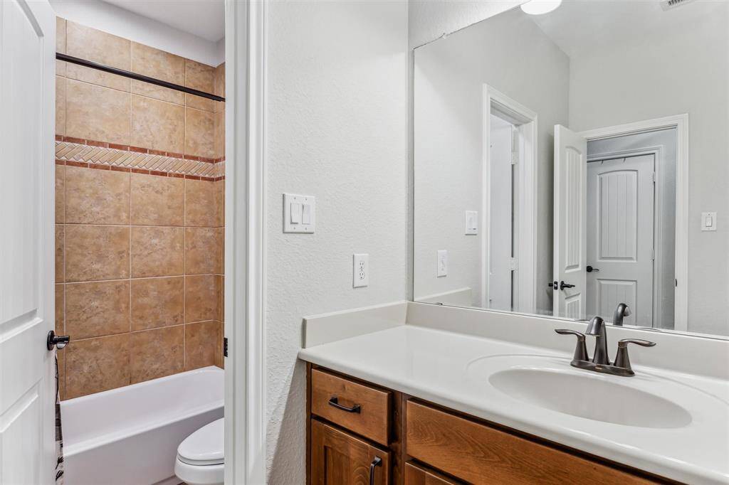 This bathroom features a clean, modern design with a single sink vanity, a large mirror, and wood cabinetry. The shower/tub combination features warm-toned tile and decorative trim