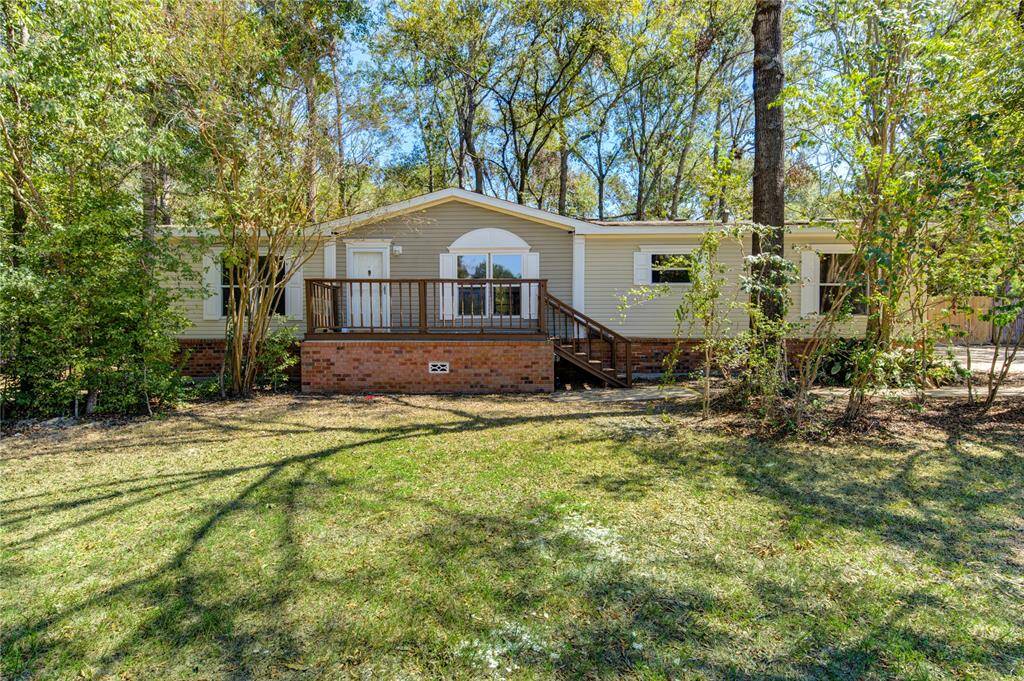 This is a single-story manufactured home with a neutral color palette, featuring a welcoming front deck, brick skirting, and nestled among mature trees for added privacy.