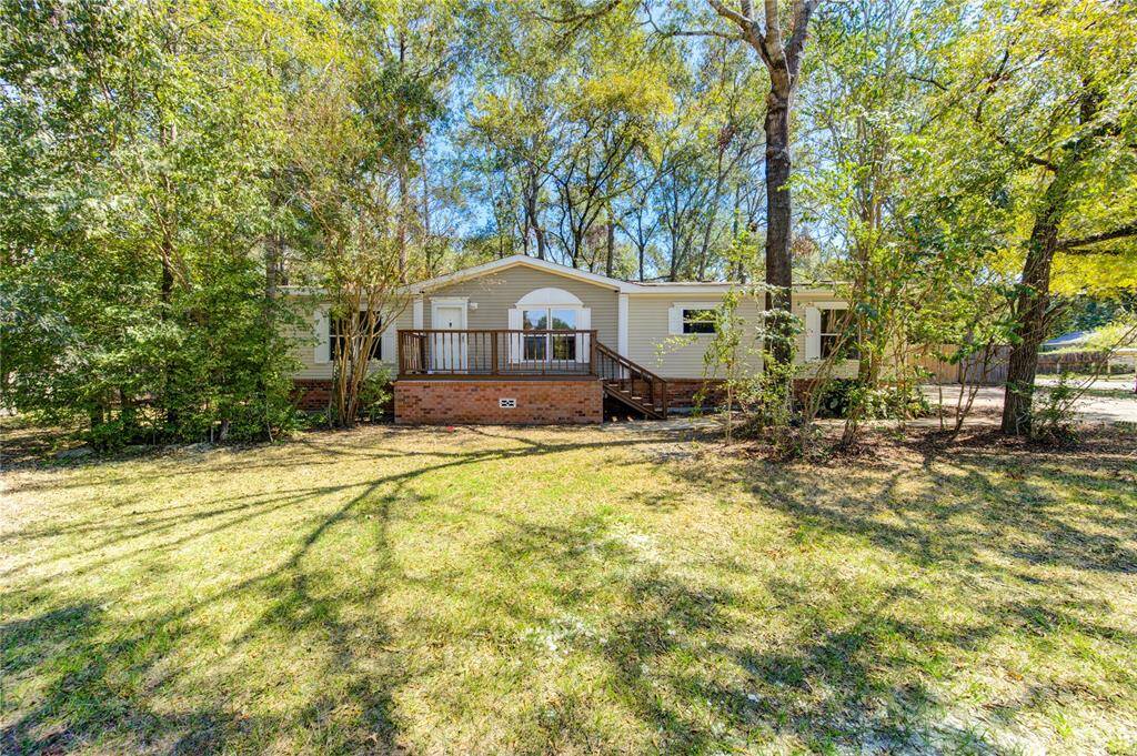 This is a single-story manufactured home with a sizable front deck, surrounded by mature trees on a sunny lot with ample yard space.