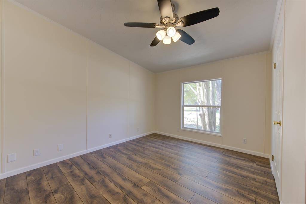 This is a bright, empty room featuring wood flooring, a ceiling fan with lighting, neutral-colored walls, and a window providing natural light.
