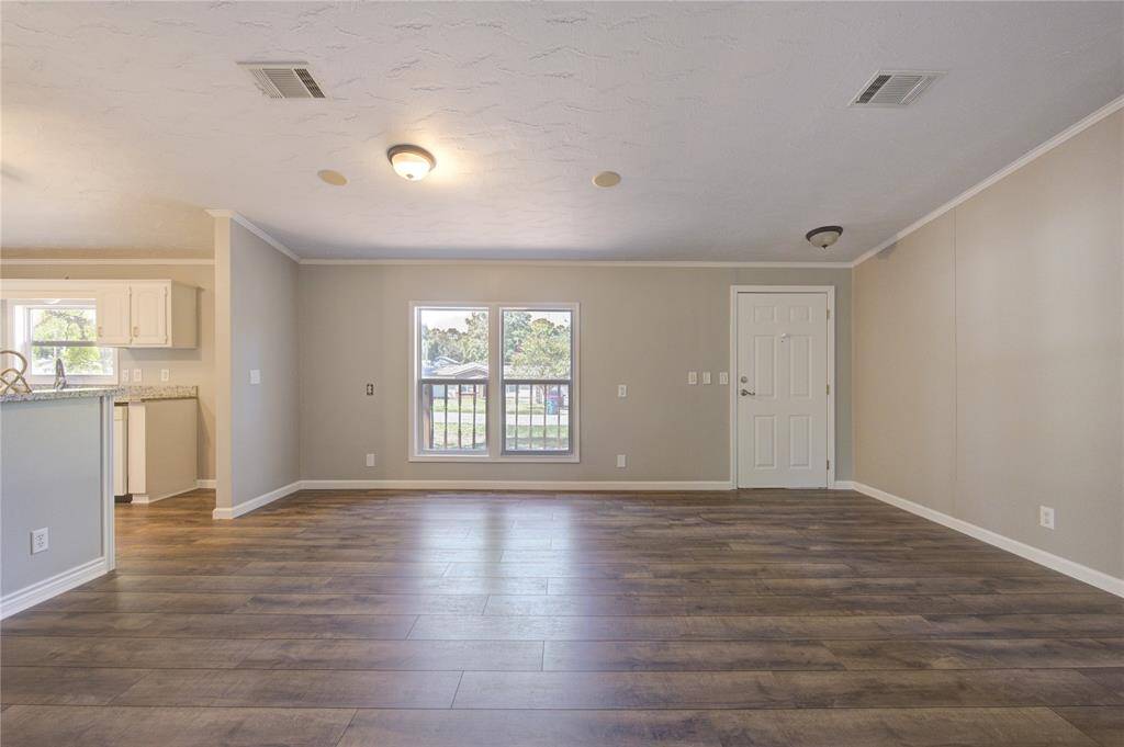 The photo showcases a spacious, open-concept living area with contemporary wood flooring, leading to an adjoining kitchen with white cabinetry. Natural light streams in through the glass doors that offer a view of the outside. The room is finished with a neutral color palette, providing a versatile backdrop for personalization.
