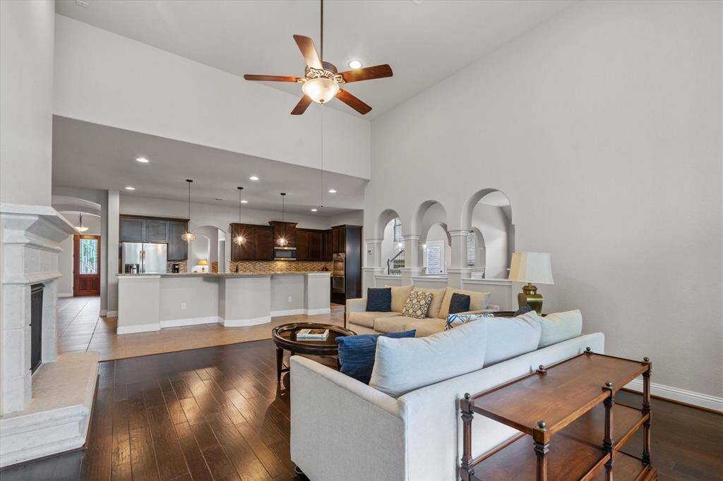 Spacious living area with high ceilings and a ceiling fan, featuring an open layout that connects to a modern kitchen with a long island. Arched openings and elegant finishes enhance the room's appeal, while hardwood floors add warmth and style.