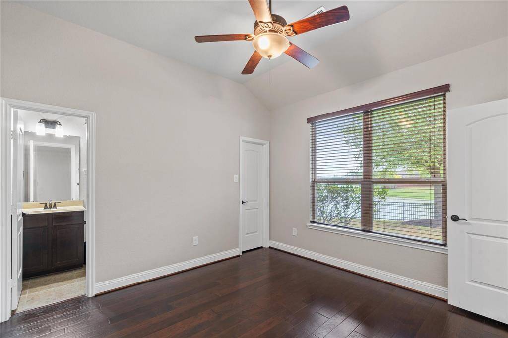 This room features hardwood floors, a ceiling fan, and a large window with blinds offering a view of the outdoors. It includes an en-suite bathroom with a modern sink and dark cabinetry, providing convenience and style.
